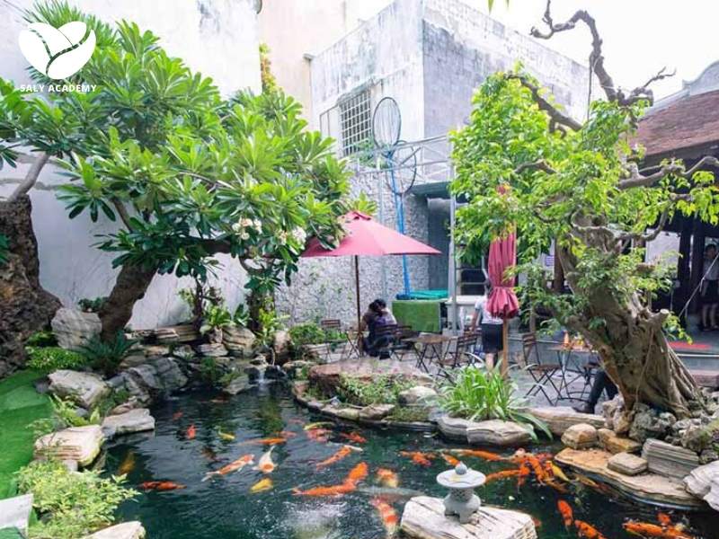 Không gian xanh mát tại một quán trà sân vườn với cây xanh hồ cá và bàn ghế gỗ tự nhiên